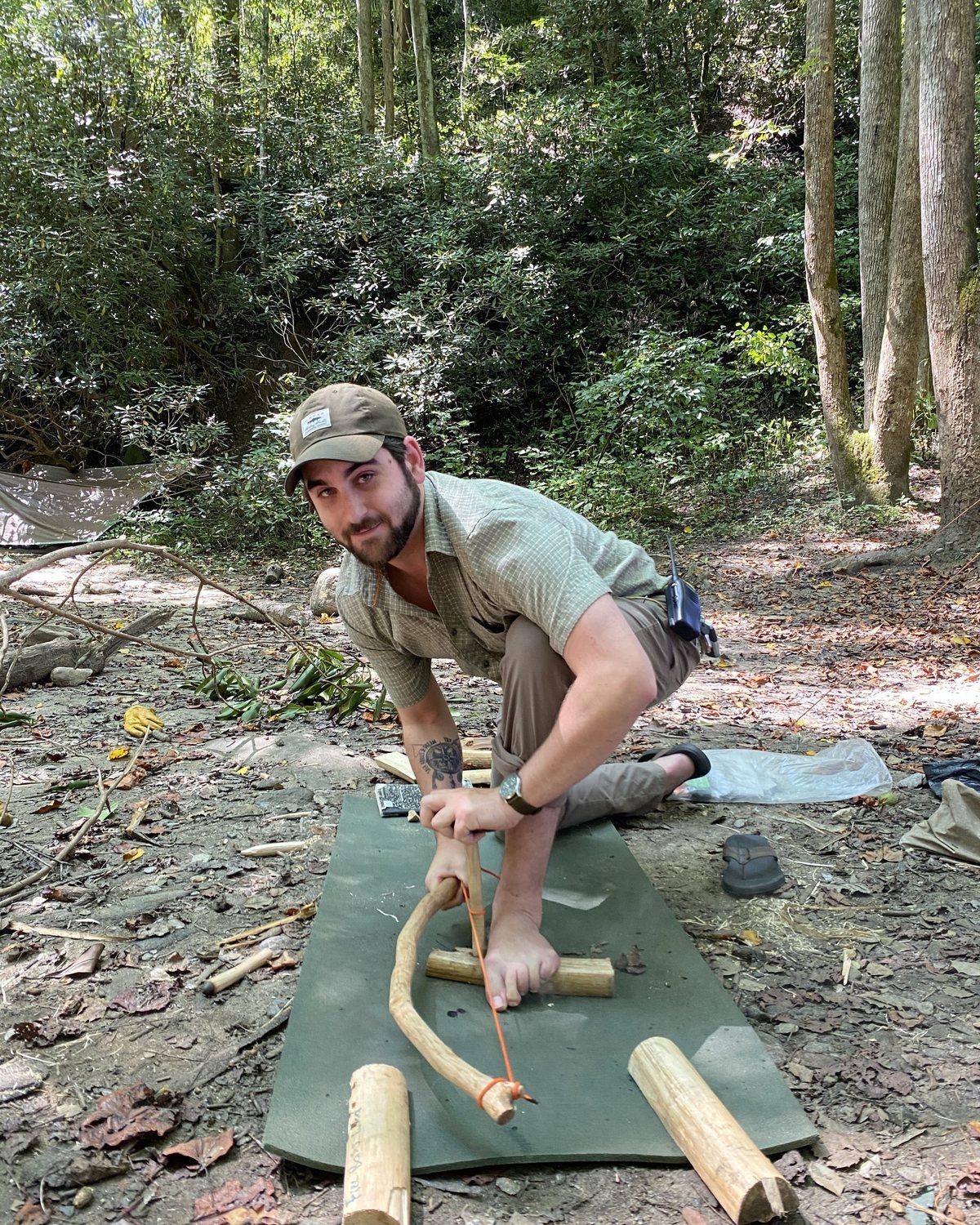 Josh Gorelick crouched on a mat in the woods, working a bow drill to start a fire, looking up at the camera.